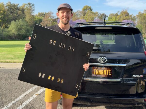 A man stands in a parking lot holding a large Trunk Mate Sleeping Platform, ready to load into his black Toyota RAV4 with a custom “TRNKMTE” plate—weekend adventures made easy.