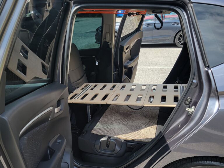 Wooden Trunk Mate Sleeping Platform set up over the back seat area of a car, shown from the open passenger side door—ready to crash wherever adventure takes you.