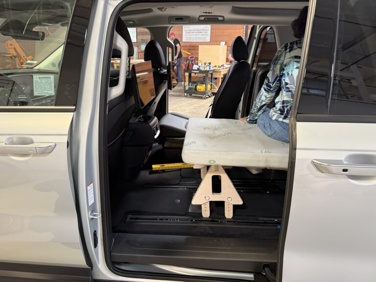 A person relaxes comfortably on a Trunk Mate Sleeping Platform set up as a bench in the backseat area of a van, with camping gear visible just outside the open sliding door.