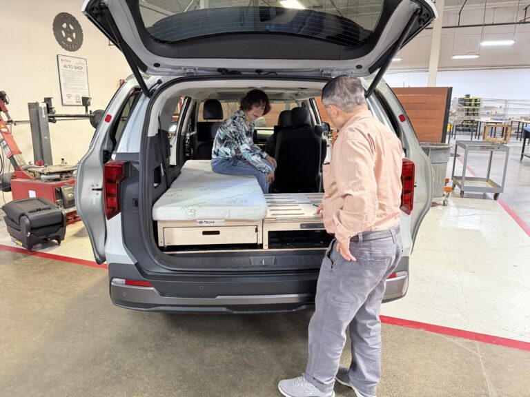 Two people check out a Trunk Mate Sleeping platform with built-in storage drawers, set up in the back of an SUV inside a workshop.