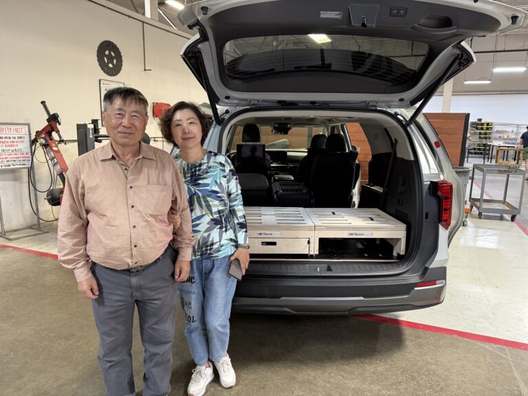 Two friends next to an open SUV, showing off their Trunk Mate Sleeping Platform setup with wooden storage crates in the back—perfect for adventure-ready organization in any space.