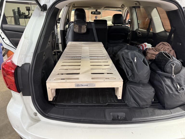 A sturdy wooden Trunk Mate sleeping platform with sleek slats set up in the back of an SUV, packed and ready for adventure with black duffel bags and travel gear arranged alongside.