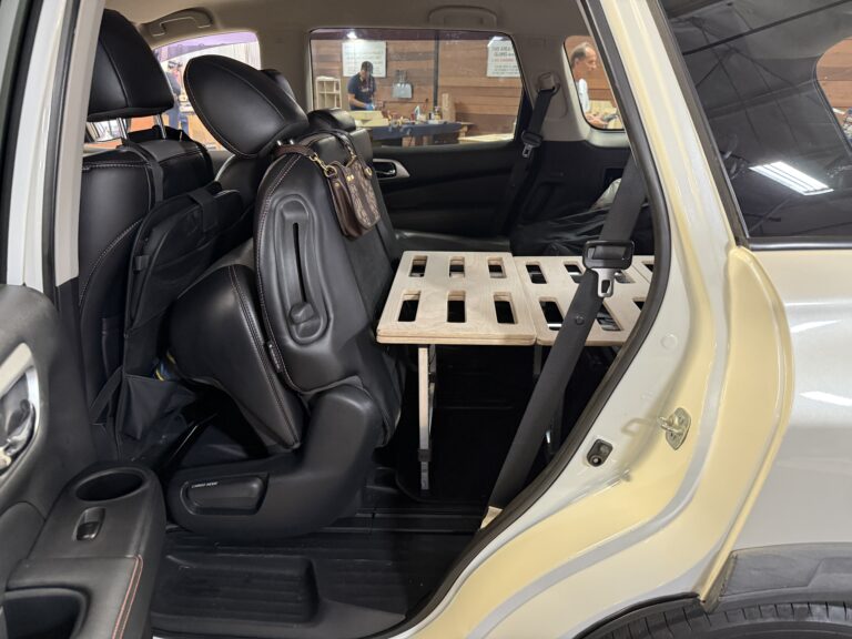 The backseat of a 2019 Nissan Pathfinder Rock Creek with a Trunk Mate wooden slatted sleeping platform set up behind the passenger seat, ready for campout vibes; people and picnic tables can be seen outside the open trunk.