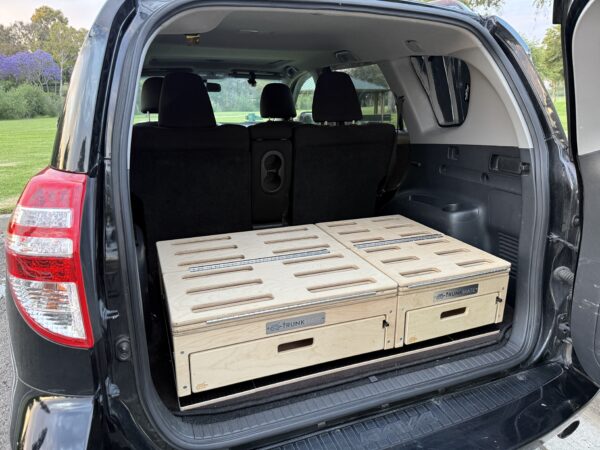 A Trunk Mate Sleeping Platform installed in an SUV trunk with the rear seats up, showing off slotted wood panels and spacious pull-out drawers for easy gear storage.