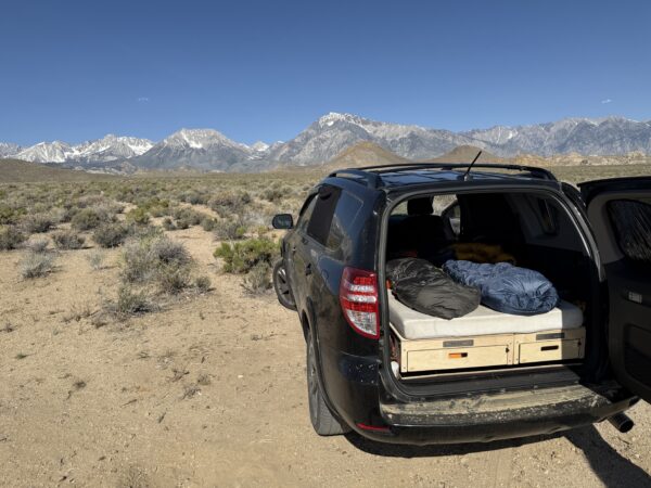 A black SUV with its rear door open shows off a comfy Trunk Mate sleeping platform, all set up for adventure. Parked on open plains with mountains and blue sky behind, it's ready for you to relax under the stars.