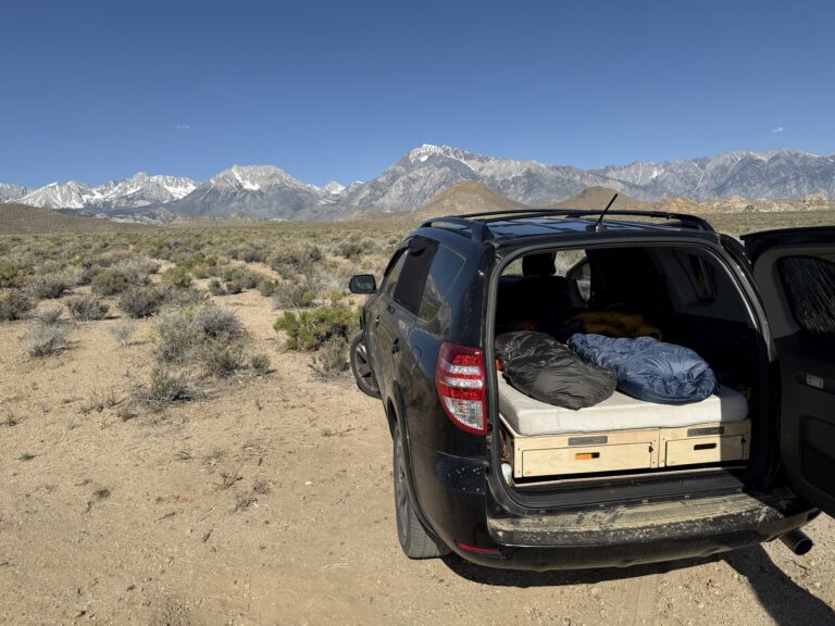 A black SUV with its rear door open shows off a comfy Trunk Mate sleeping platform, all set up for adventure. Parked on open plains with mountains and blue sky behind, it's ready for you to relax under the stars.
