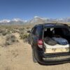 A black SUV with its rear door open shows off a comfy Trunk Mate sleeping platform, all set up for adventure. Parked on open plains with mountains and blue sky behind, it's ready for you to relax under the stars.