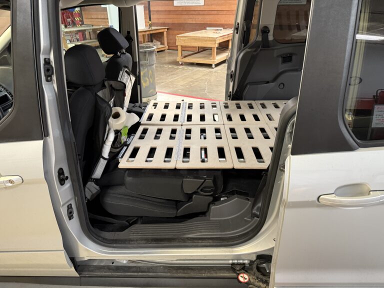 A sturdy wooden slatted Trunk Mate Sleeping platform set up in the back of a car, resting above folded seats with road trip essentials like paper towels nearby—ready for spontaneous adventure or comfy camping.