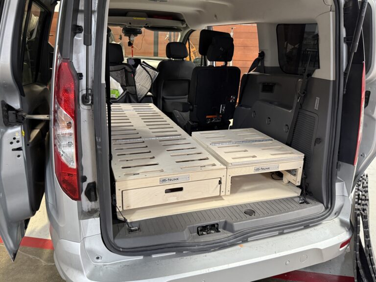 The back of a van with the doors open, featuring a Trunk Mate wooden slatted sleeping platform bed and roomy storage drawers underneath—your ticket to organized, comfy road trips.