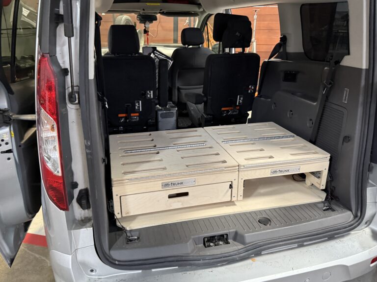 The back of a van with both rear doors open, featuring a Trunk Mate Sleeping Platform—custom wooden storage drawers neatly installed in the cargo space for organized camping and easy gear access.