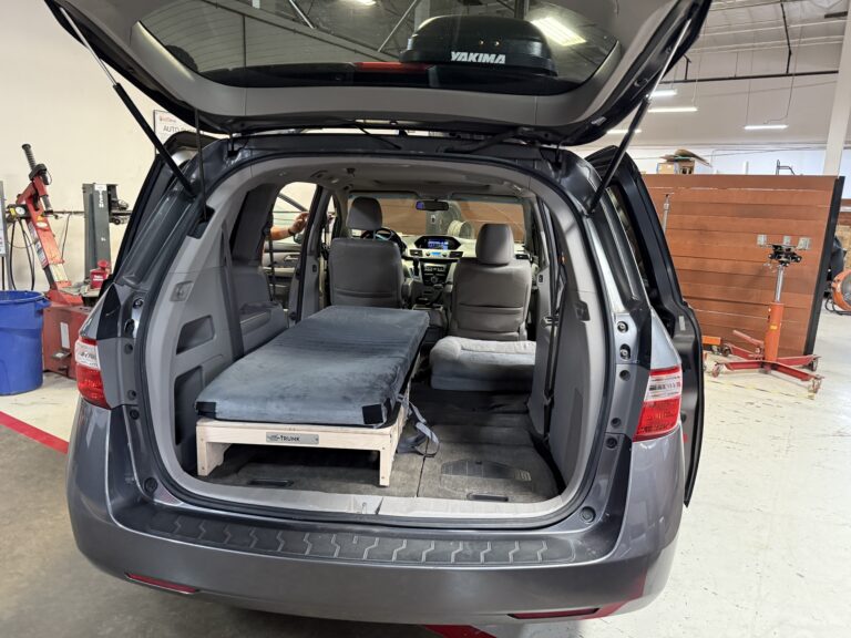 A minivan with its rear hatch open, featuring the Trunk Mate single sleeping platform and comfy mattress set up in the cargo space—perfect for easy car camping or road trips.