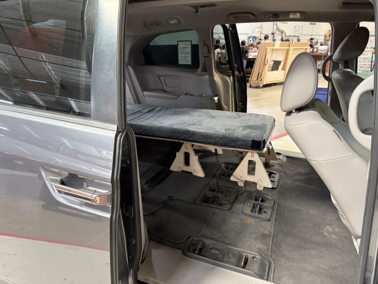 A minivan interior with seats removed, featuring a Trunk Mate collapsible sleeping platform set up like a cot—showing how easy it is to turn your van into a cozy sleep space.