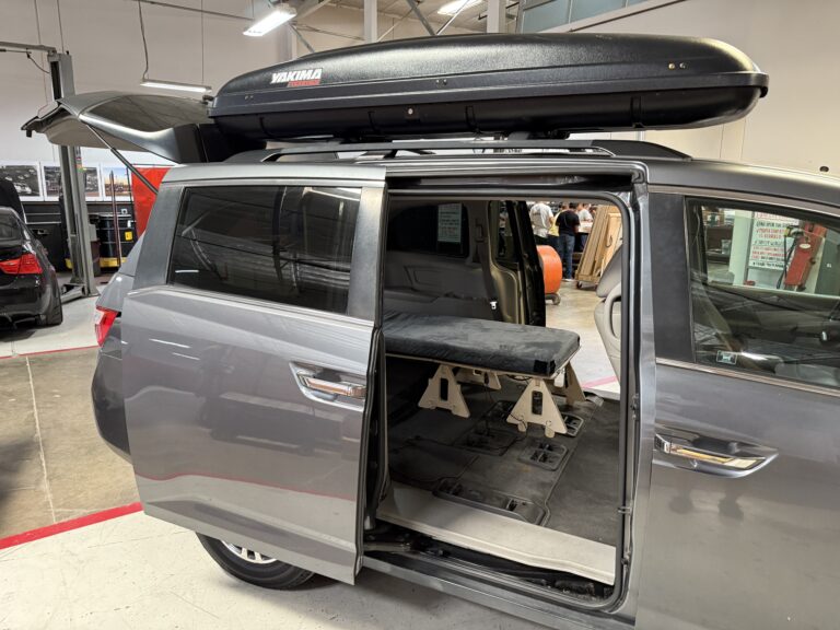 A minivan with its sliding door open reveals a Trunk Mate bench-style sleeping platform set up inside, ready for adventure. Up top, a Yakima cargo box provides extra gear storage for the trip.