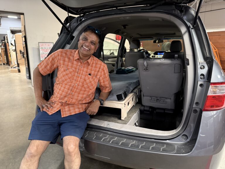 A man in an orange plaid shirt and blue shorts relaxes at the open trunk of an SUV, showing off a Trunk Mate custom sleeping platform set up in the back.