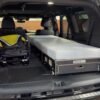The back of an SUV loaded for adventure—a folded stroller sits on the left, while a comfy Trunk Mate sleeping platform with mattress is set up on the right, showing just how much gear and sleep space you can fit in your cargo area.