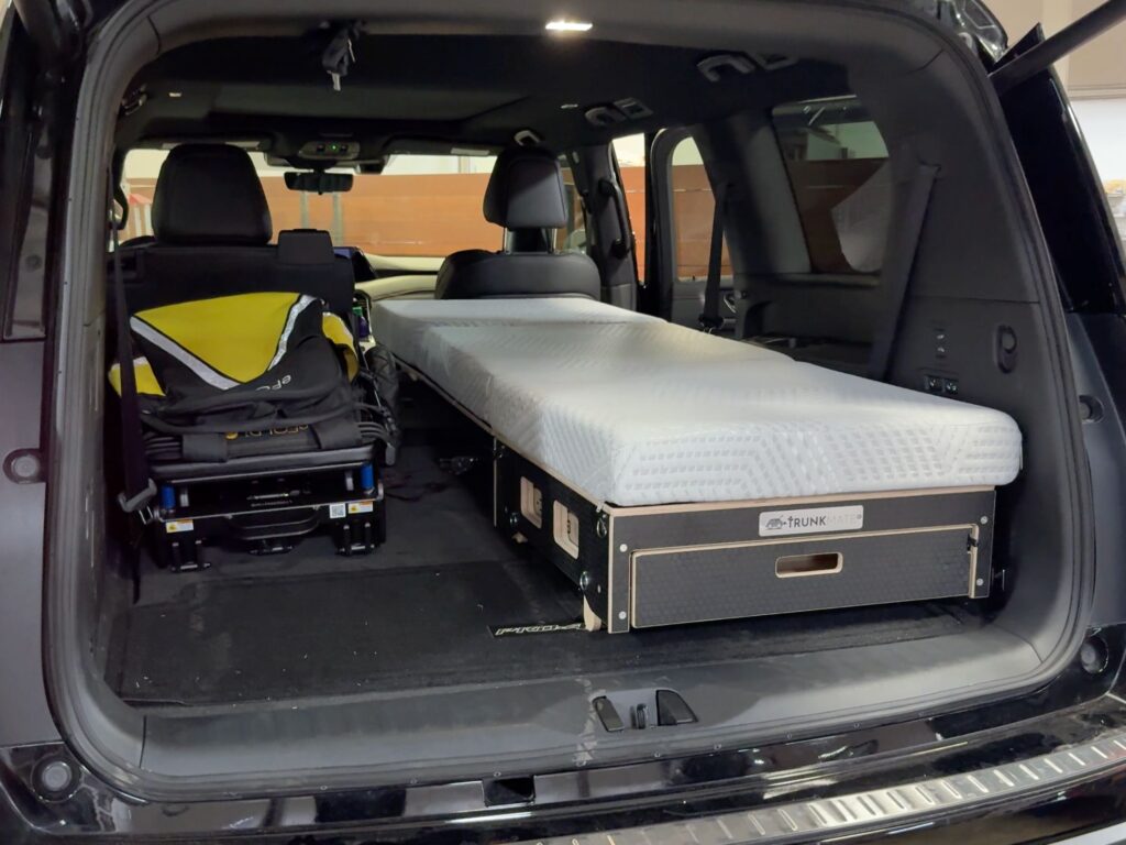 The back of an SUV loaded for adventure—a folded stroller sits on the left, while a comfy Trunk Mate sleeping platform with mattress is set up on the right, showing just how much gear and sleep space you can fit in your cargo area.