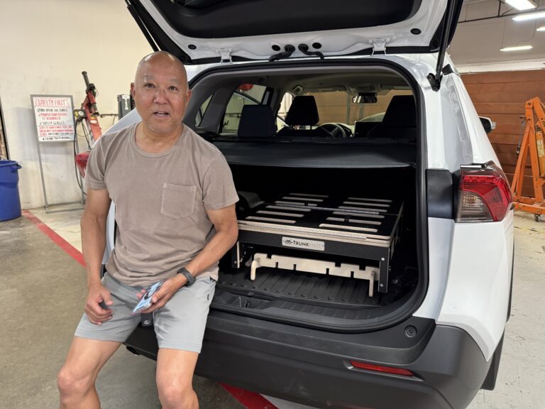 A man relaxes on the open tailgate of his SUV, showing off the Trunk Mate Sleeping Platform neatly installed in the trunk—ready for adventure and organized storage.