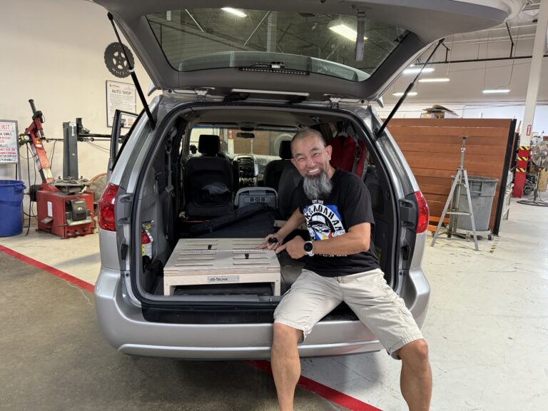 A smiling man sits at the open trunk of a minivan in a workshop, holding a cardboard box while relaxing on a Trunk Mate Sleeping Platform, ready for the next adventure.