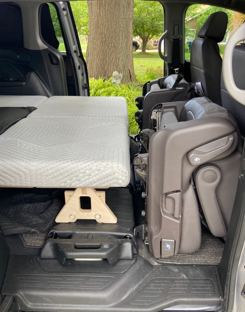 Back seats folded forward with a Trunk Mate Sleeping Platform and mattress set up inside the car, creating a cozy sleeping space with green trees visible through the windows.