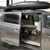 Gray minivan with sliding door and rear hatch open, roof box on top, and Trunk Mate sleeping platform set up inside.