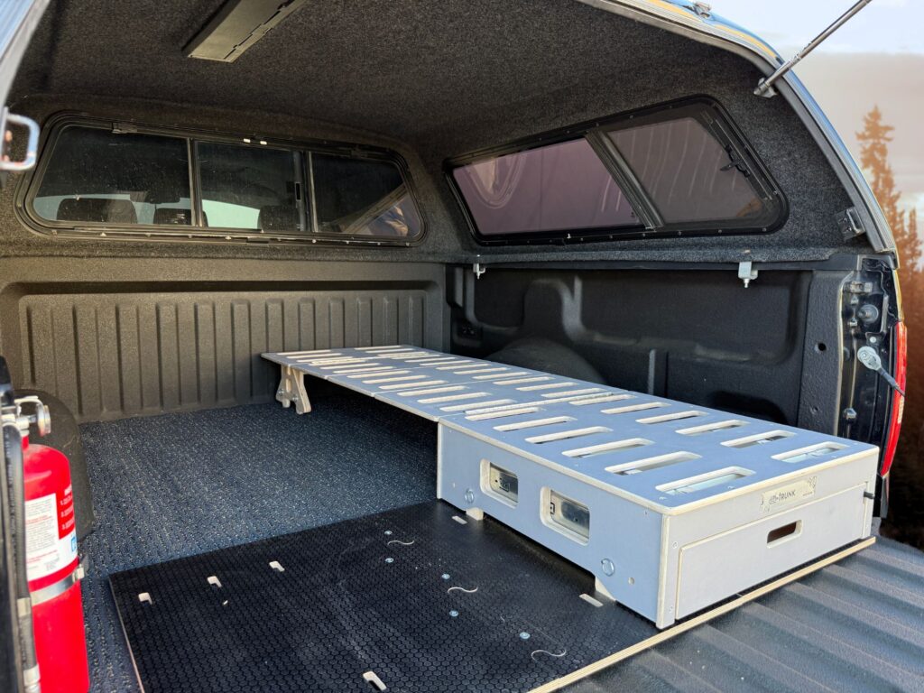 A pickup truck bed with a canopy, outfitted with a sturdy metal Trunk Mate Sleeping platform and spacious storage drawer underneath; a red fire extinguisher is conveniently mounted by the tailgate for added peace of mind on your adventures.